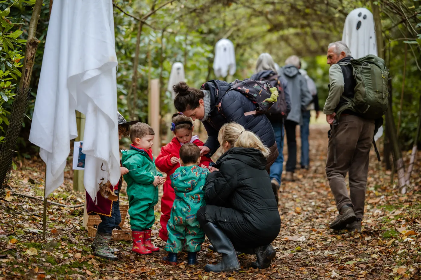 Toddlers in waterproofs and an adult crouching beside them on a leaf-covered path lined with hanging ghosts, with walkers behind
