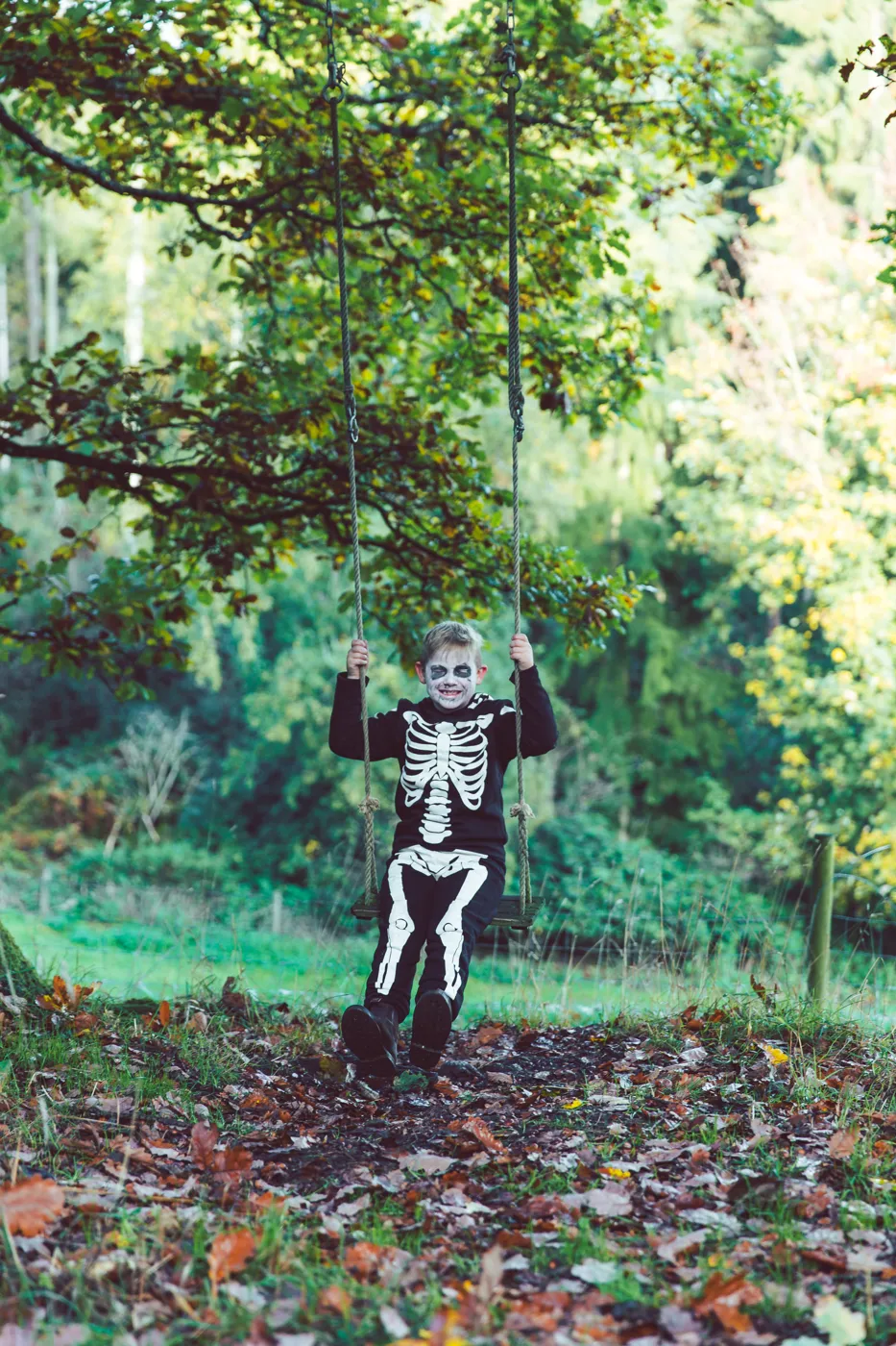 A child in a skeleton costume and face paint sitting on a rope swing under a tree