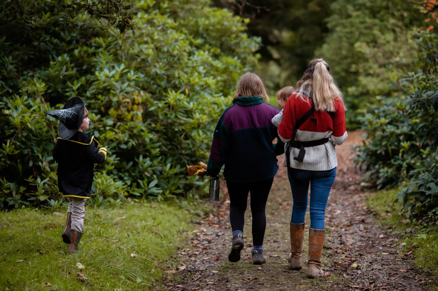 A child in a witch's hat walking beside a woman and a girl carrying a toddler along a woodland path between rhododendrons