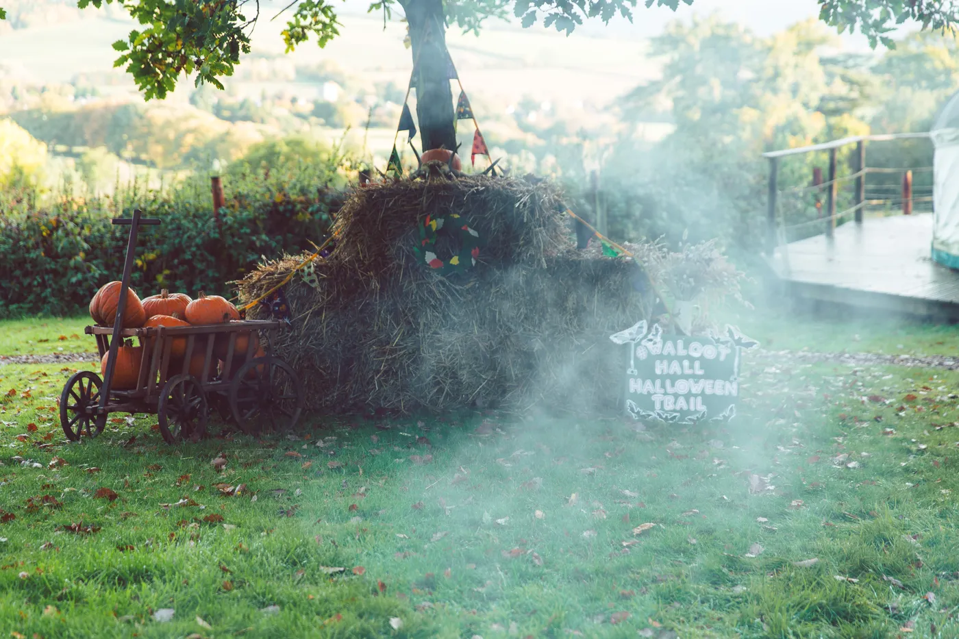 Hay bales, a cart of pumpkins and a chalkboard sign reading Walcot Hall Halloween Trail, with mist drifting across the grass
